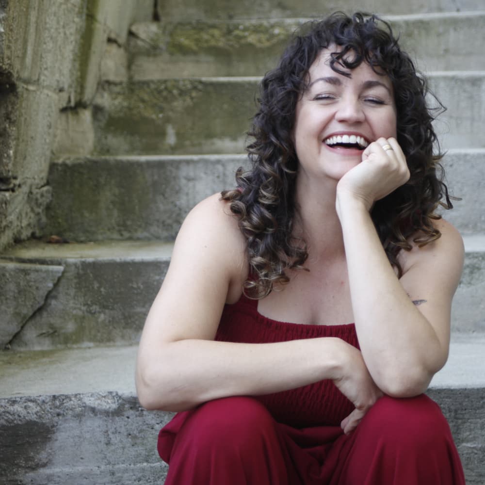Eve Sturges wears a red sleeveless jumpsuit, laughing at the camera, seated on cement stairs.